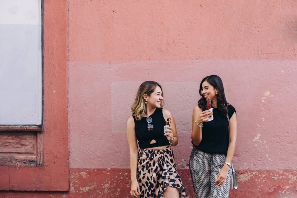 visit a new city new year goal idea. two friends smiling against a big pink wall on a building while eating a treat